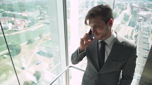 Young Businessman in a Suit Talks on a Mobile Phone and Rides in a Panoramic Elevator in a alt