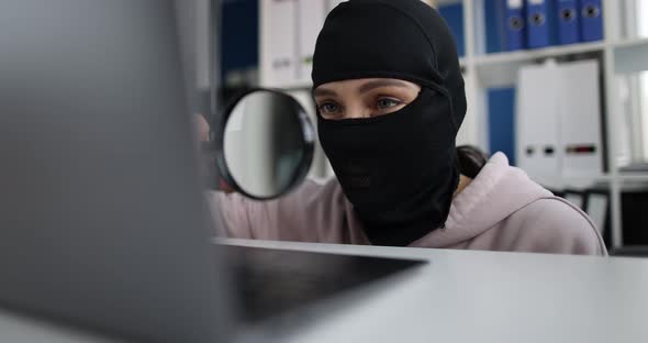 Cybercriminal in Black Balaclava Looks Through Magnifying Glass Laptop Monitor alt