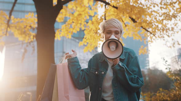 Young Caucasian Woman with Shopping Bags Announcing Sales Season with Loudpeaker in the City Park in alt