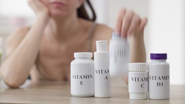 Blurred Thoughtful Woman Sitting at Table with Row of Vitamin Bottle Choosing Pills alt
