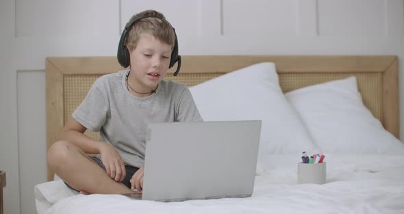 Little Boy Is Sitting on Bed in Home and Chatting Online By Notebook, Talking at Microphone of alt