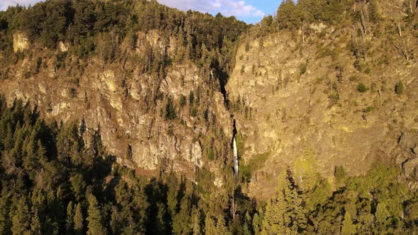 Aerial flying over Corbata Blanca thin waterfall between pine tree forest mountains at golden hour, alt