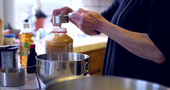 An elderly woman measuring ingredients and pouring them into a metal mixing bowl while baking a vega alt