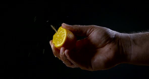 Man Is Compressing Half of Orange Between Fingers, Juice Is Splashing in Sides, Black Background alt