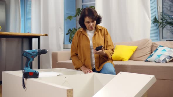Senior Woman Assembling Furniture at Home and Calling Carpenter alt