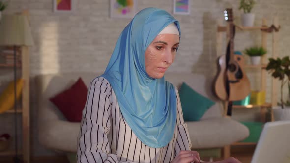 Portrait Young Muslim Woman in a Hijab with a Scar From a Burn on Her Face Works at a Laptop alt