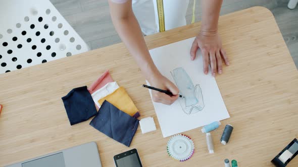 Closeup of Female Designer's Hands Drawing Sketches of New Trendy Clothes on Paper on Table alt