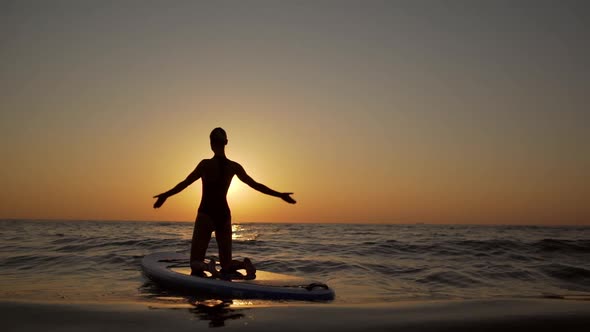 Young Woman Doing Knee Yoga Asana on Supboard on Waves with Raised Hands in Slowmotion alt