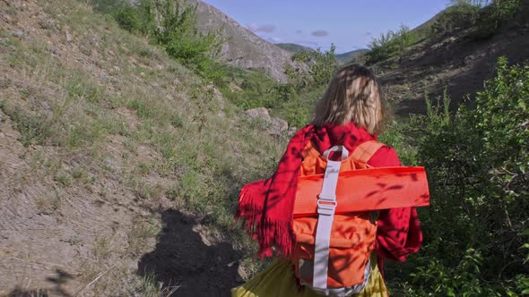 Young Woman with a Red Backpack Goes on a Mountain Road in a Hike The Camera Is Smoothly Watching alt