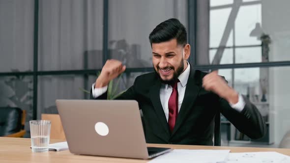 Excited Indian or Arabian Businessman in Suit Sits in a Modern Office Looking at Laptop Happy By alt