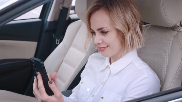 Beautiful Young Woman Sitting in a Car on Drivers Seat and Using Smartphone alt