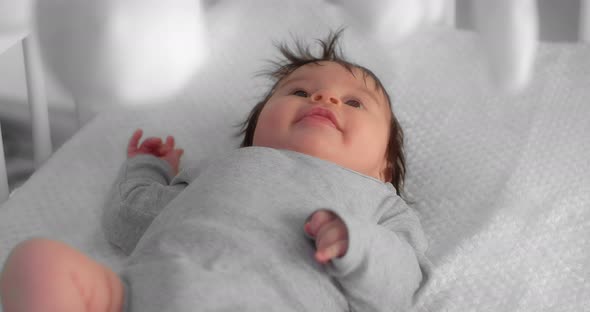Newborn Baby in a Gray Bodysuit is Lying in a Crib and Looking at a Child Mobile