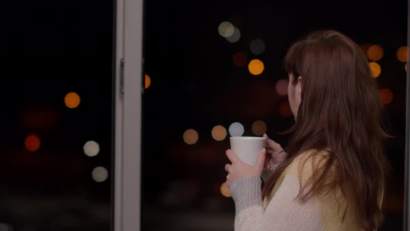 Woman Near the Window in the Evening Drinks Hot Coffee By Passing Car Lights