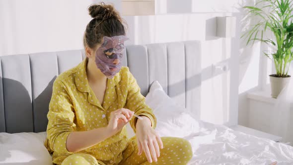 Woman in Pajamas with Sheet Mask Applies Cream on Hands