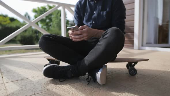 Young man using a smartphone while sitting at longboard on terrace of house alt