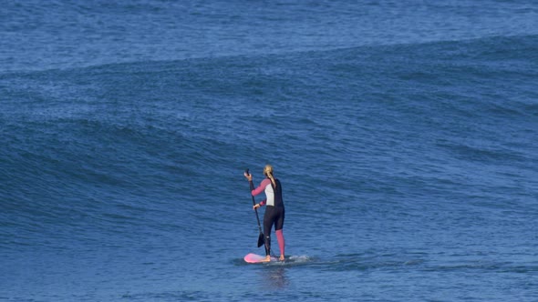 A woman rides an sup stand up paddleboard while surfing on a pink surfboard. alt