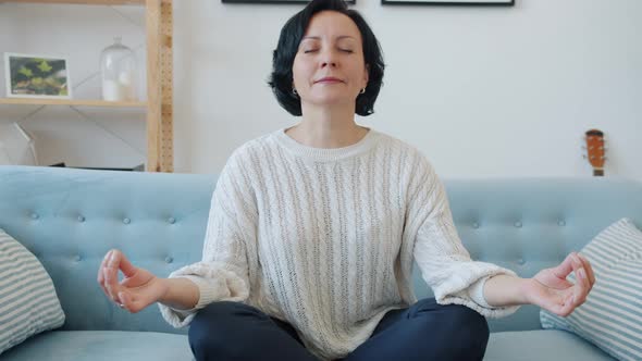 Mature Lady Sitting on Couch in Lotus Pose and Meditating with Eyes Closed Indoors at Home alt