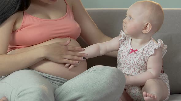 Toddler and expecting female sitting on sofa, child touching belly with hand alt
