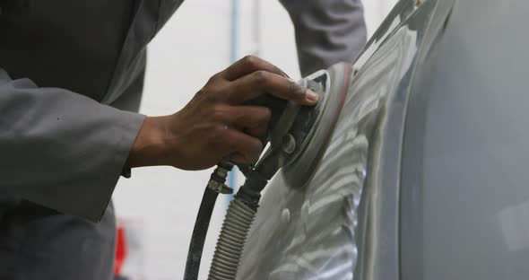 African American male car mechanic wearing a face mask and polishing a side of a car with a grinder alt