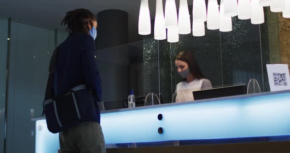 Mixed race man and female receptionist wearing face masks at hotel reception desk alt