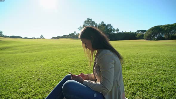 Side View of Young Girl Using Smartphone on a Grassy Field with the Sun Shining in the Background