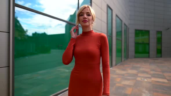 a Blonde in a Red Dress Walks Along a Modern Building and Looks at Her Reflection in the Large Green alt