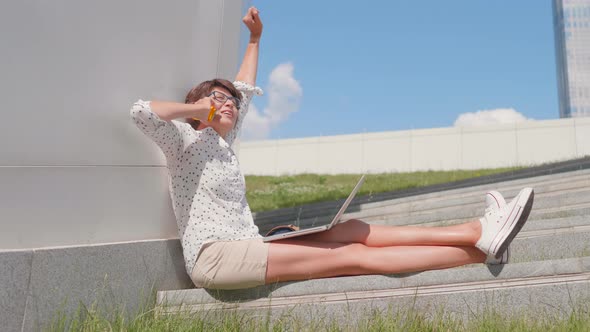 Woman in Eyeglasses Works with Laptop Ant Talks By Smartphone in Urban Park alt