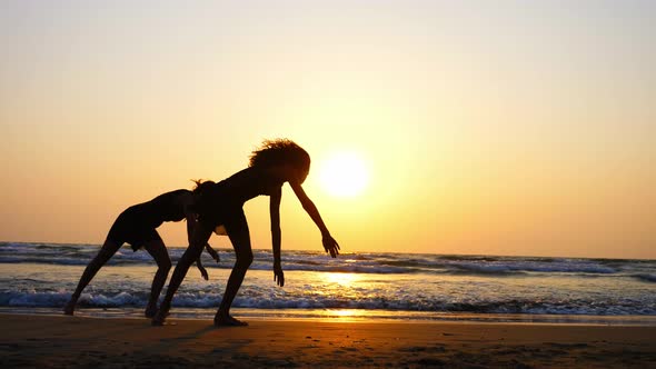 Silhouette of Sporty Young Women Practicing Acrobatic Element on the Beach alt