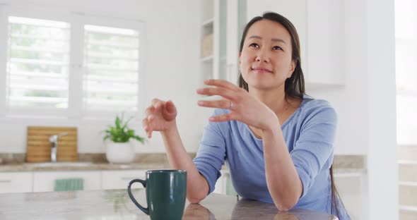 Happy asian woman sitting in kitchen and drinking coffee alt