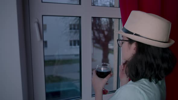 Girl in a Hat with a Glass of Wine Pulls Back the Curtain and Looks Attentively Out the Window Young