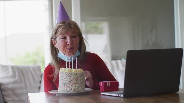 Senior woman blowing cake while having a video chat on laptop at home alt