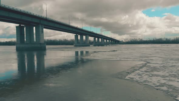 Grey Bridge Over Wide River with Ice Against Dense Forest alt