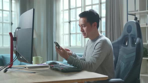Asian Man Using Mobile Phone While Using Desktop Computer For Working At Home alt