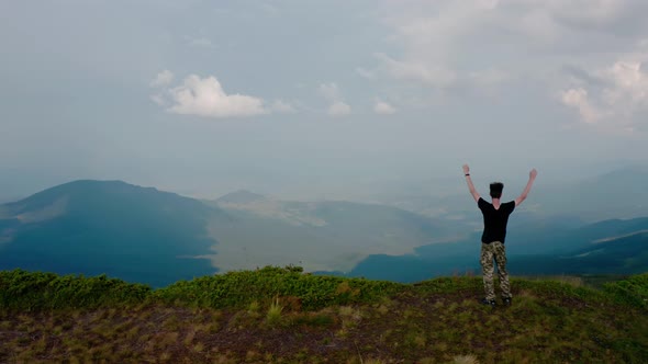 Aerial Drone View. Flying Around Young Man Standing on Top of the Mountain at Sunset alt