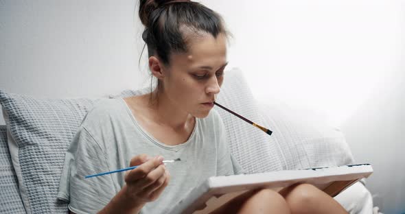 Young Woman Artist Painting Canvas in Her Living Room with Paint Brush in Hand alt