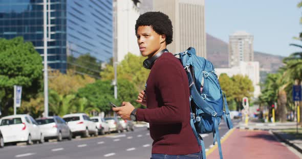 African american man in city, using smartphone, wearing headphones and backpack crossing street alt