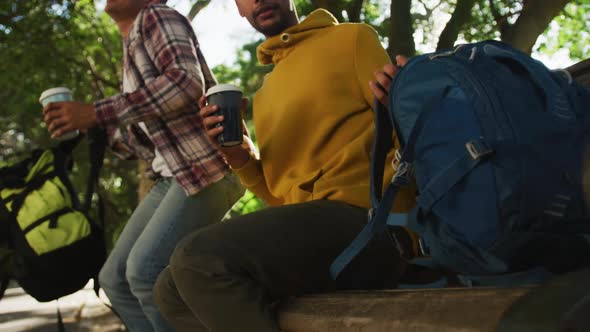 Two happy mixed race male friends drinking coffee in park with backpacks alt