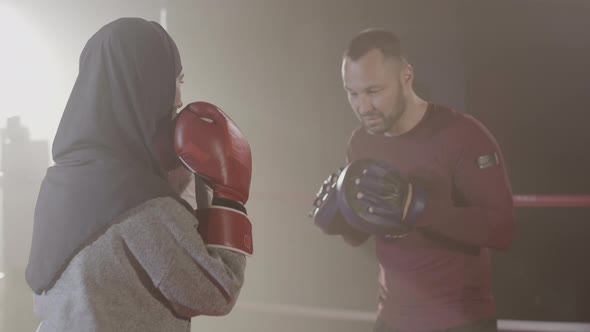 Back View of Young Woman in Hijab Punching Training Gloves of Coach in Backlight Fog. Confident alt