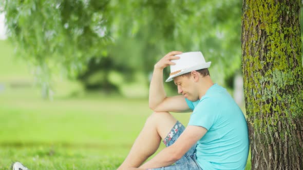 Young man is sad while sitting under a tree in a city park, blurred background alt