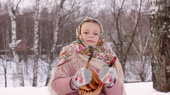 Cute girl in a traditional Russian headscarf and mittens playing on spoons on winter forest backgrou alt