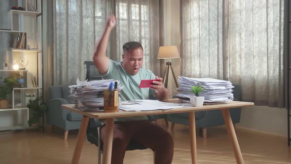 Happy Fat Asian Man Celebrating Winning Game On Smartphone After Working With Documents alt