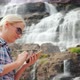A Traveler Uses a Smartphone Against the Background of a Waterfall Tvindefossen in Norway - VideoHive Item for Sale
