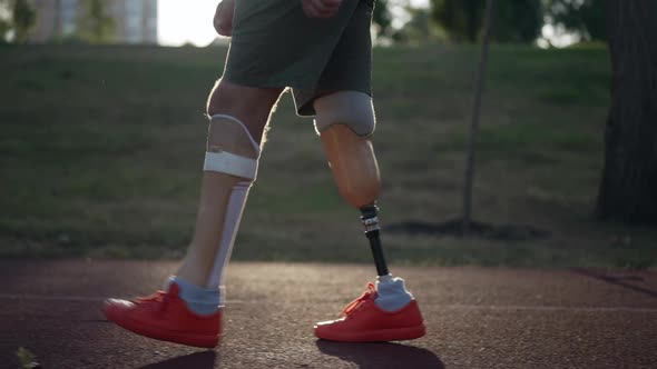 Side View Prosthetic Legs of Male Amputee Walking in Sunrays in Slow Motion on Red Sports Track alt