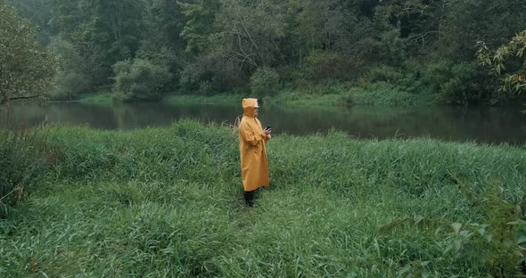 Worker in Helmet and Raincoat Walks to River on Grass Man Has Phone in His Hand