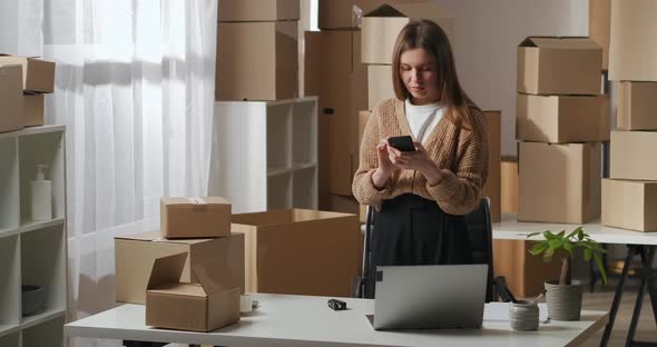 Young Woman in Her Working Flat with Boxes Using Cell Phone for Communicating By Messenger alt