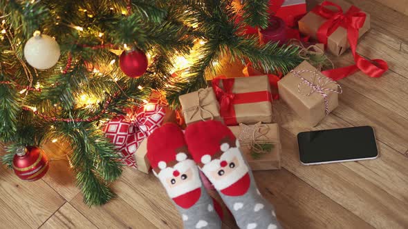 Feet in Funny Christmas Socks Santa Face Wooden Floor Under Fir Tree Gift Boxes alt