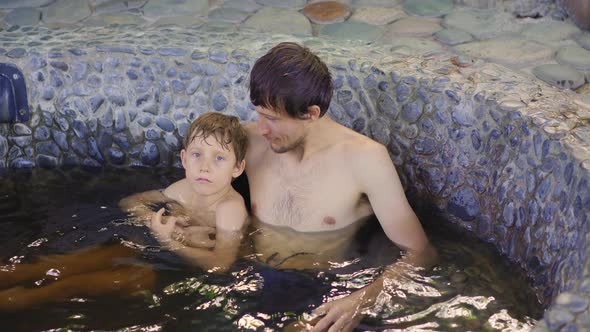 Young Man and His Son Relax in a Stone Bath Filled with Healing Herbal Infusions alt