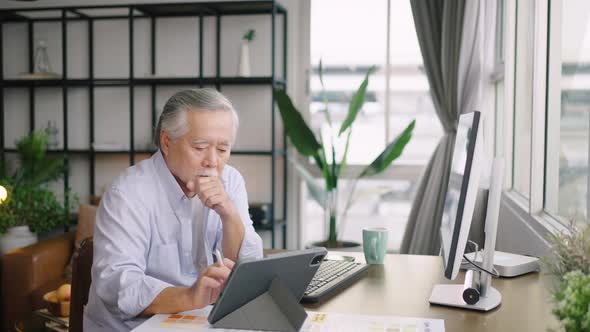 Asian senior mature man working on computers and tablet at home and talking to customers in a busy