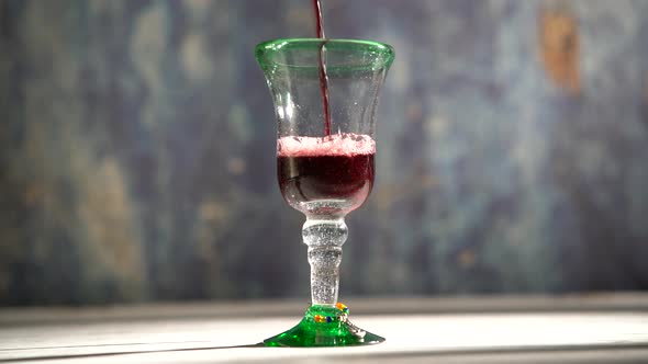 Rustic glass and painted wood table with red wine pouring into a glass with blue background. alt