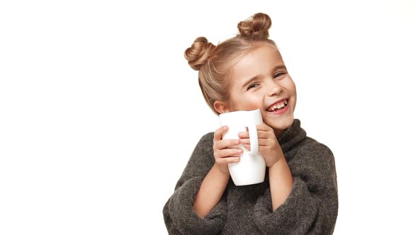 Small Beautiful Caucasian Kid Girl in Cozy Sweater Drinking From Cup on White Background and Smiling alt
